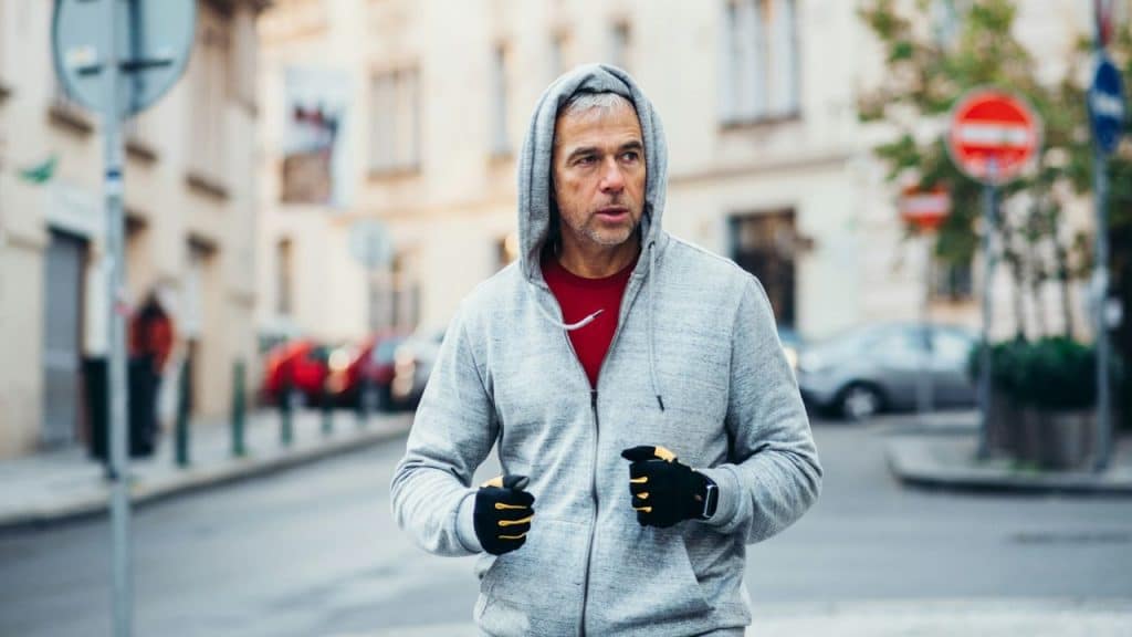 Man in a gray hooded sweatshirt and black gloves jogging on an urban street.