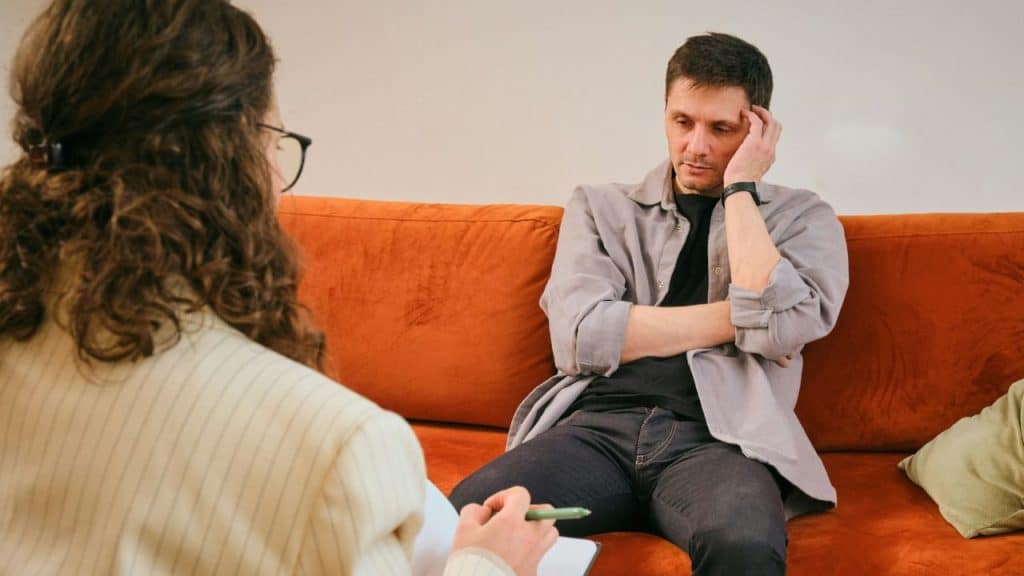 Man seated on an orange couch looking down with a hand on his head, facing a woman with a notepad.