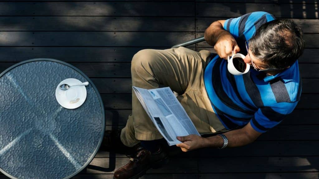 Overhead view of a man reading a newspaper and drinking coffee at an outdoor table on a dark wooden deck.