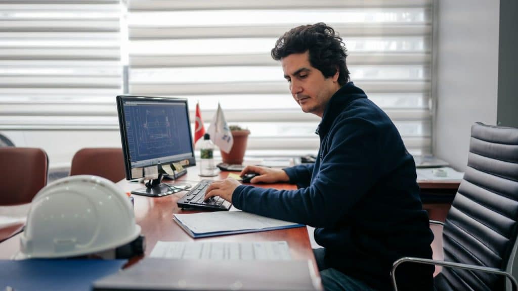 Man in a dark shirt intently working at a computer with a hard hat resting on the desk in an office.