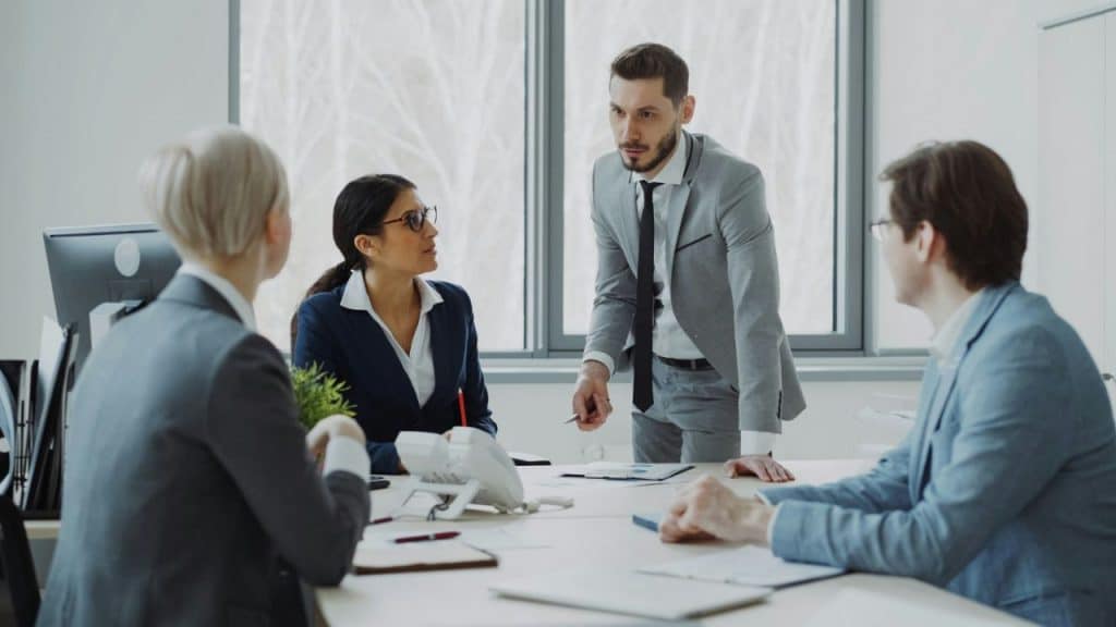 Four professionals in business attire are engaged in a serious meeting around a white conference table.