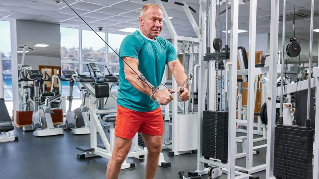 Older man in a teal shirt and red shorts using a cable machine for a chest workout in a gym.