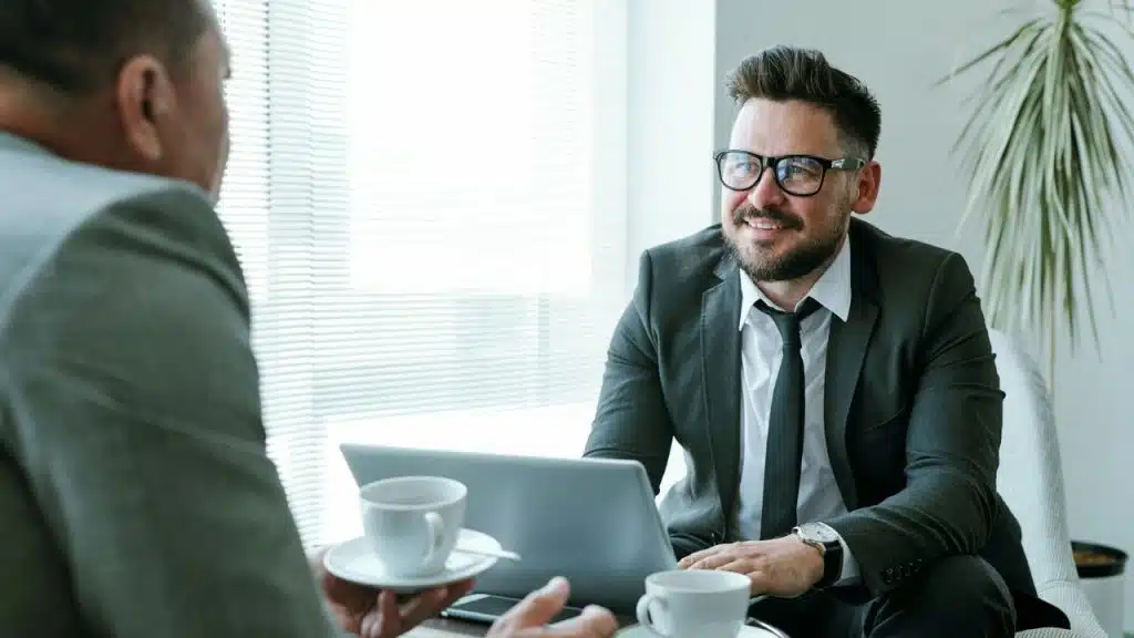 Two men in business suits talking at a table with coffee cups and a laptop near a bright window.