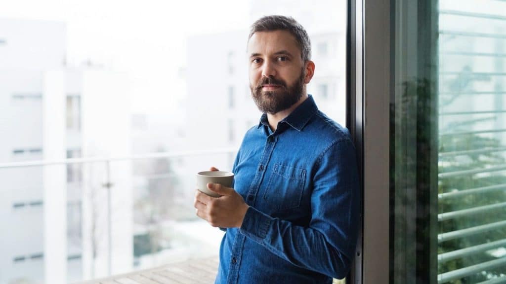 Bearded man in a blue denim shirt holding a mug while standing by a window or balcony outdoors.