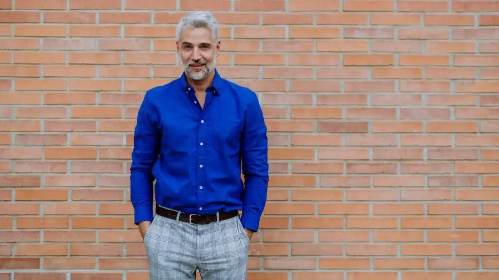 Smiling man with gray hair in a bright blue shirt and plaid pants standing against a red brick wall.