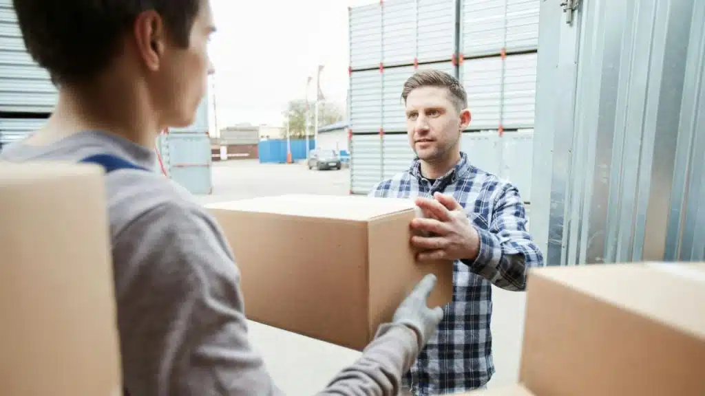 Two men handling a large cardboard box outdoors in a shipping or storage facility setting.