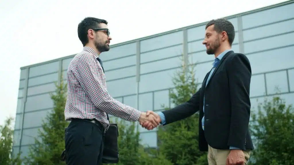 Two men in business attire are shaking hands outdoors in front of a modern glass building.