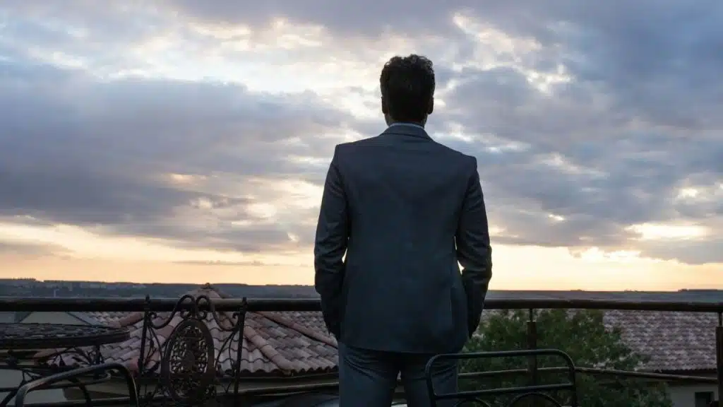 Man in a dark suit standing on a balcony, viewing a sunset over a distant landscape.