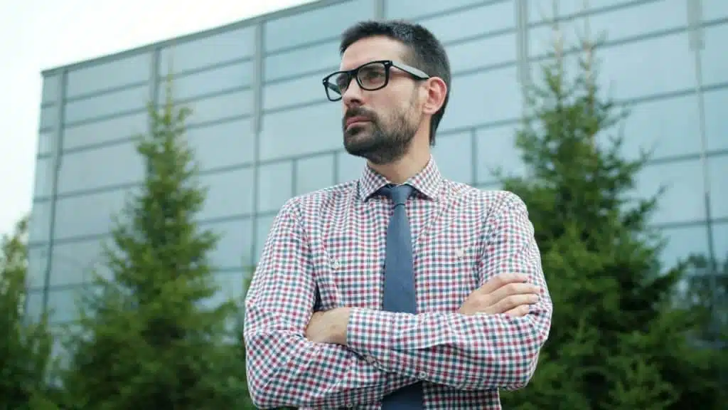 Man in a plaid shirt, tie, and glasses stands with his arms crossed in front of a modern glass building.