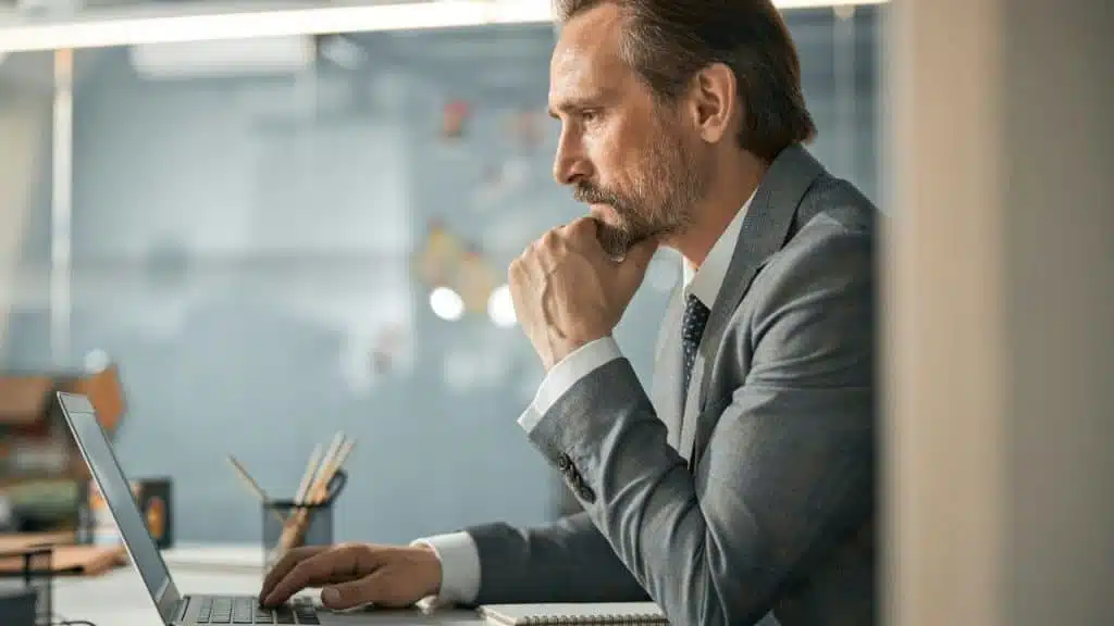 Middle-aged man in a suit with a beard is focused on a laptop with his hand on his chin in an office setting.
