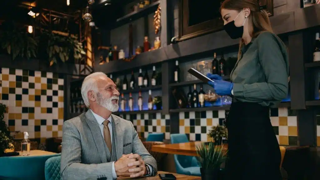 Smiling older man in a suit seated at a table talking to a masked waitress holding a tablet in a restaurant.