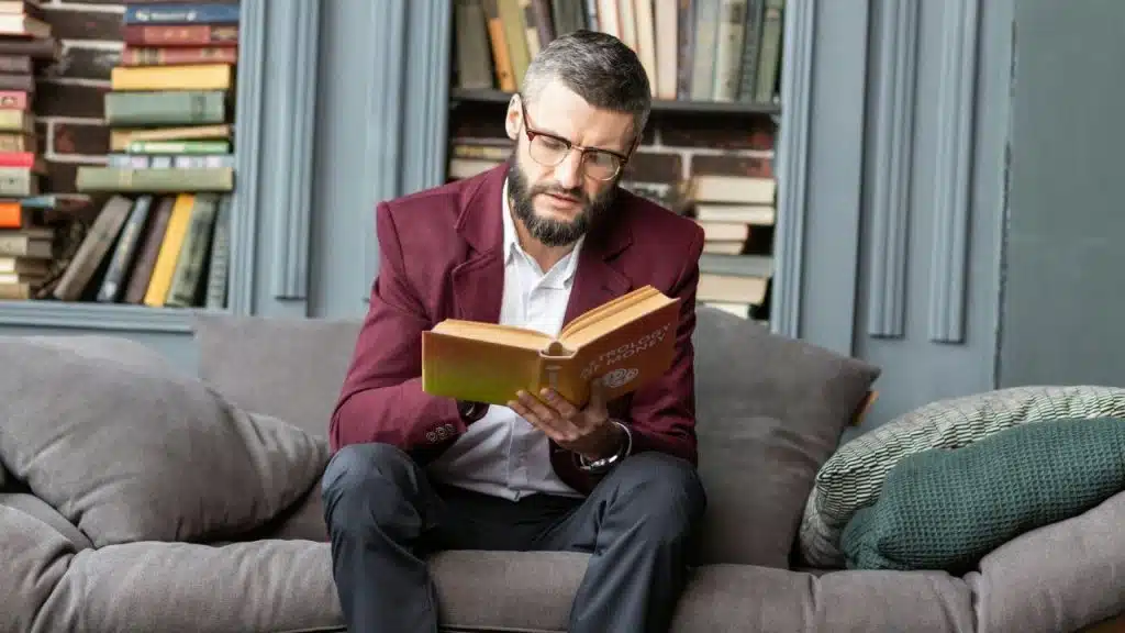 Man in a maroon jacket and glasses sitting on a couch, intently reading an open book.