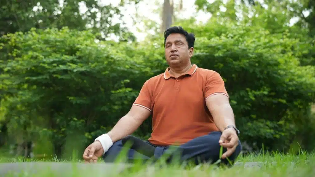 Man in an orange shirt sitting cross-legged and meditating outdoors on a grassy lawn.