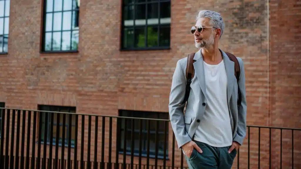 Mature man with gray hair, sunglasses, a light gray jacket, and a backpack standing by a railing near a brick building.