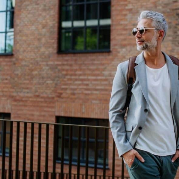 Mature man with gray hair, sunglasses, a light gray jacket, and a backpack standing by a railing near a brick building.