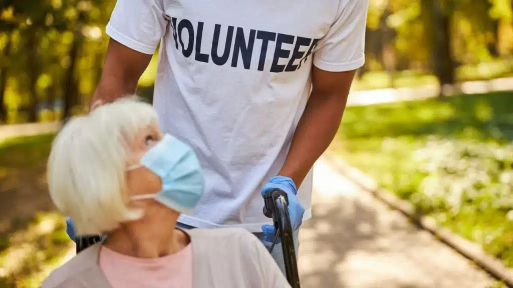 Volunteer in a white t-shirt pushing an older woman in a wheelchair wearing a face mask on an outdoor path.