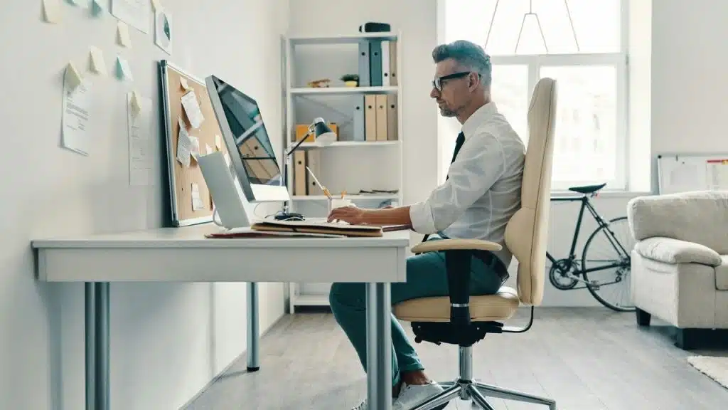 Man in a white shirt and glasses sitting in a swivel chair working at a computer desk in a bright office.
