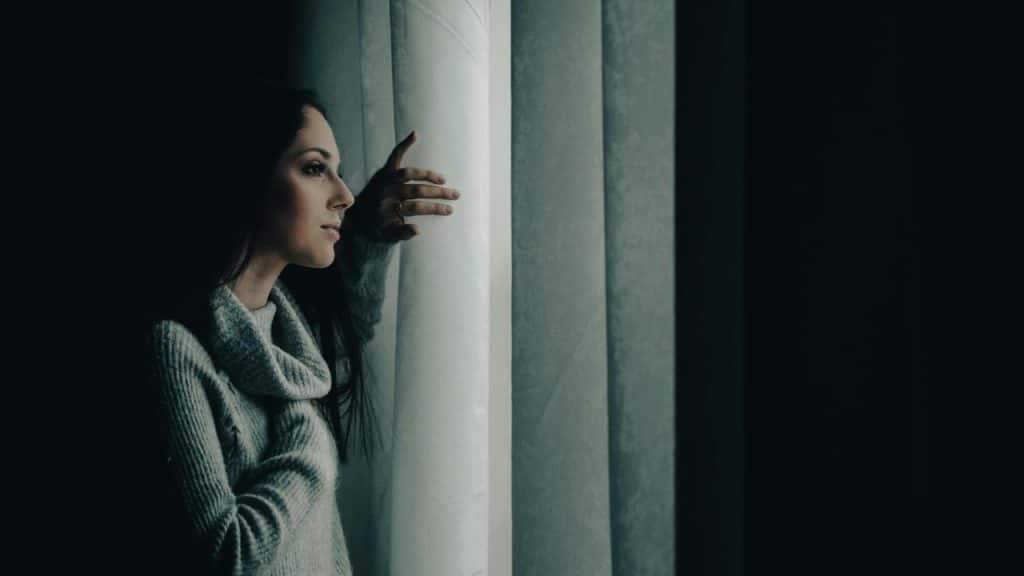 A woman looking out a window while holding the curtain.