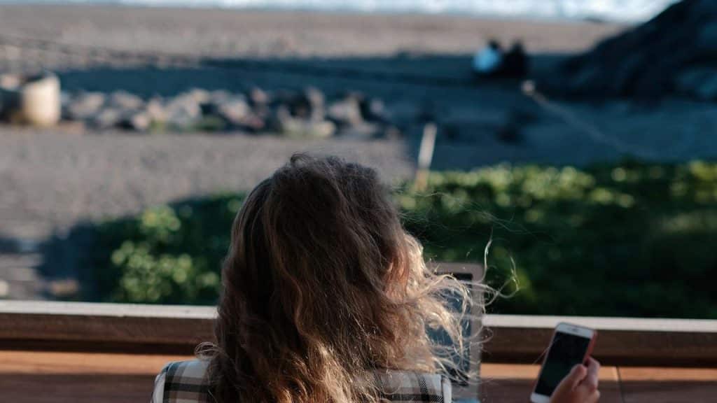 A woman sitting by the beach holding a smartphone.