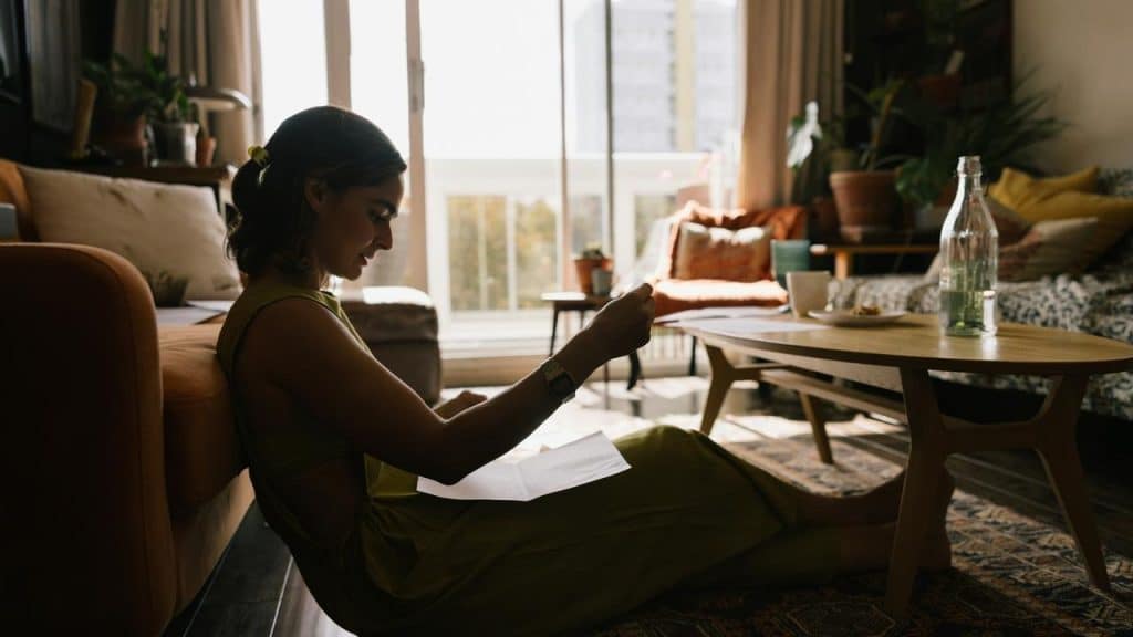 A woman sitting on the floor by a couch reading a letter in a sunlit living room.