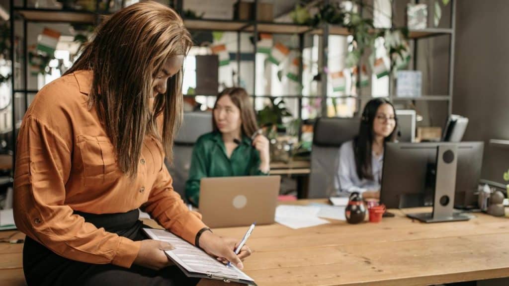 A woman writing on a clipboard while two colleagues work at their desks in an office.