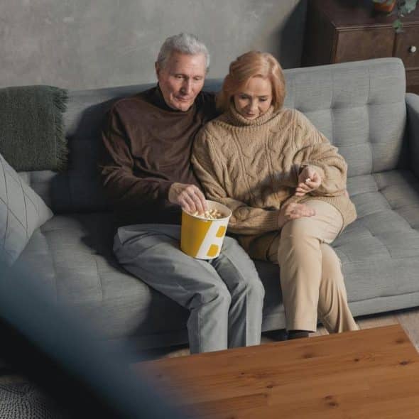 A senior couple sitting on a couch eating popcorn together.