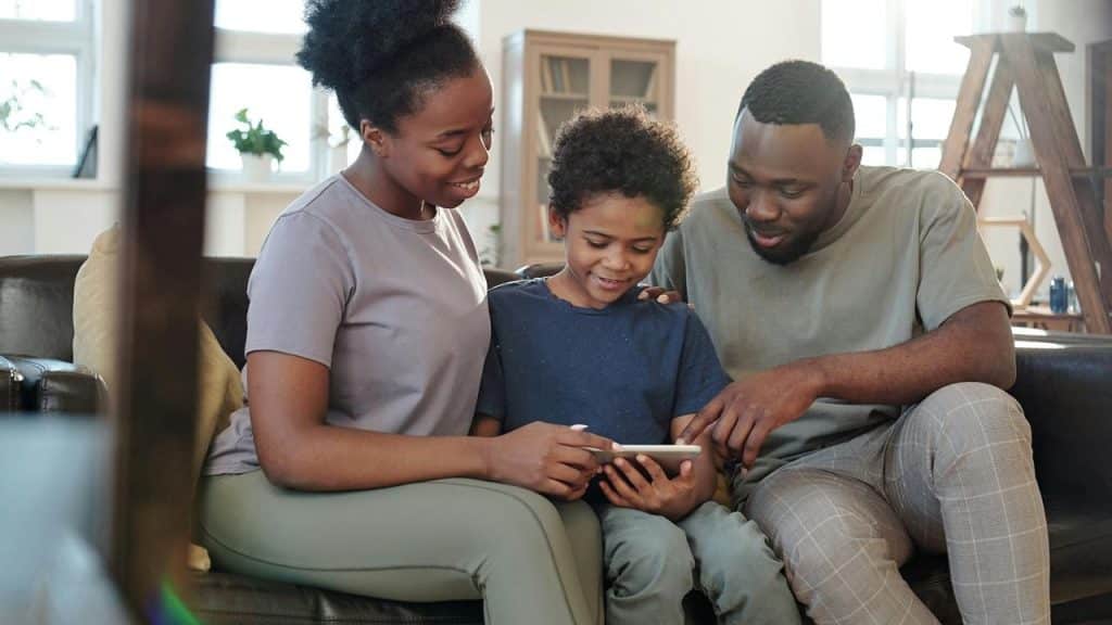 A family sitting together on a couch, smiling while looking at a tablet.