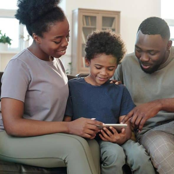 A family sitting together on a couch, smiling while looking at a tablet.
