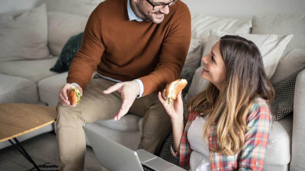 A couple working on a laptop while eating sandwiches.