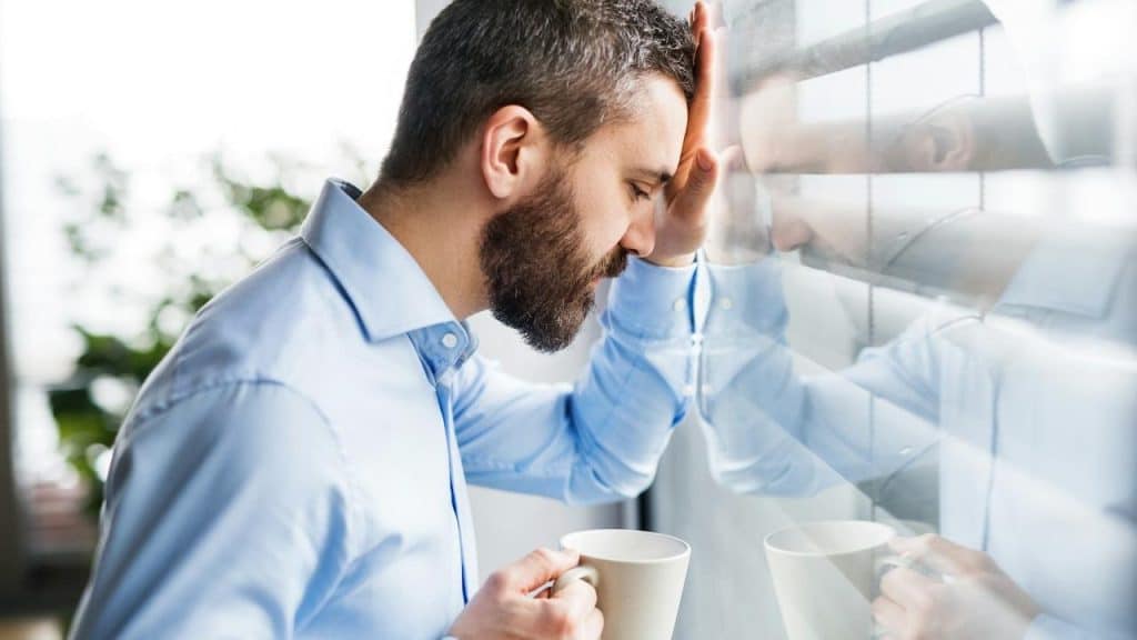 A man leaning his head on a glass window while holding a coffee cup.