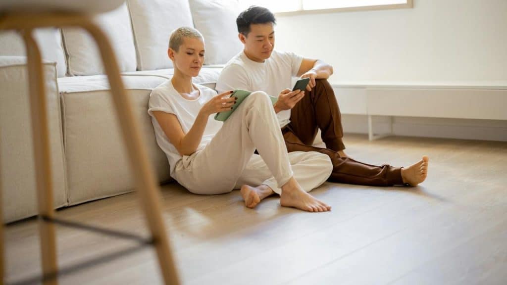 A couple sitting on the floor by a couch while looking at their phones.