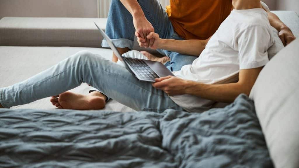 A person sitting on a couch with a laptop on his lap and reaching for the hand of another person.