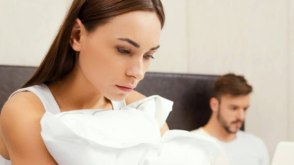 A woman looking sad and holding a pillow while an oblivious man works on his laptop in the background.