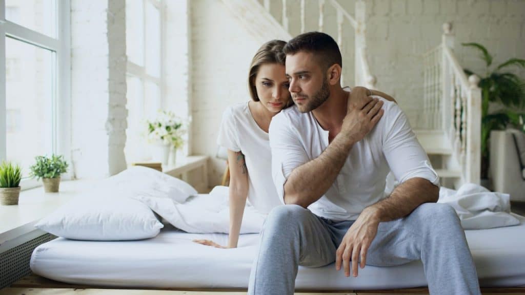 A man looking distressed while a woman holds him from behind and sitting on a bed.