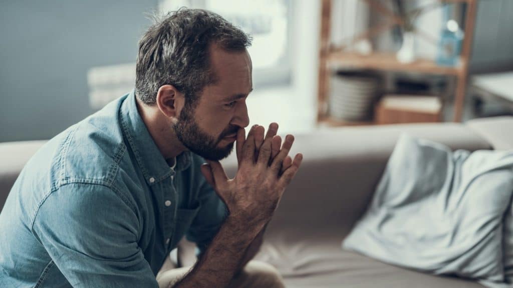 A bearded man holding clasped fingers to his mouth and looking depressed in his living room.
