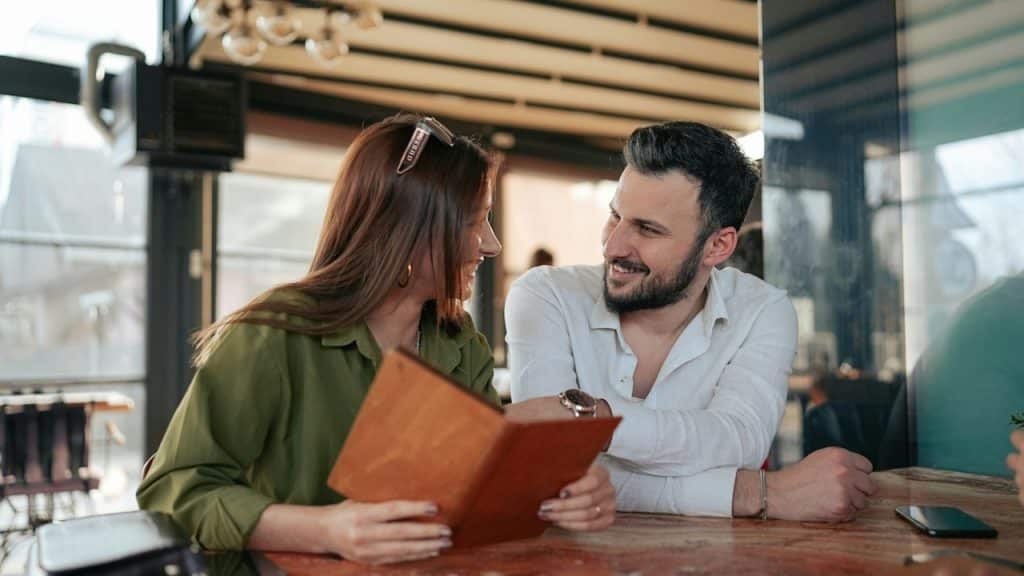 A smiling man and woman looking at each other across a table while holding a menu in a well-lit cafe or restaurant.