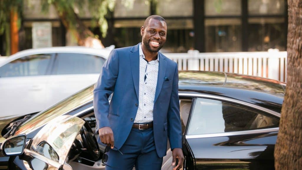 Smiling man in a blue suit standing by the open door of a black luxury car.