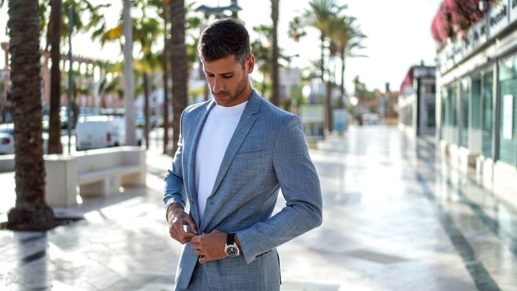 Handsome man in a light blue suit and white t-shirt adjusting his jacket on a sunny, palm-tree-lined walkway.