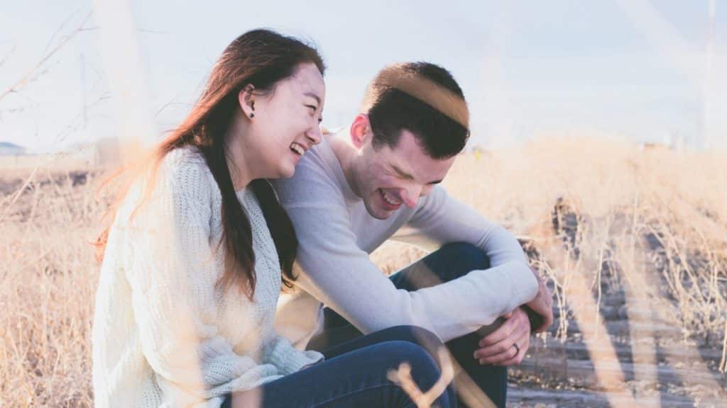 Young couple sitting outdoors laughing together in tall, dry grass.