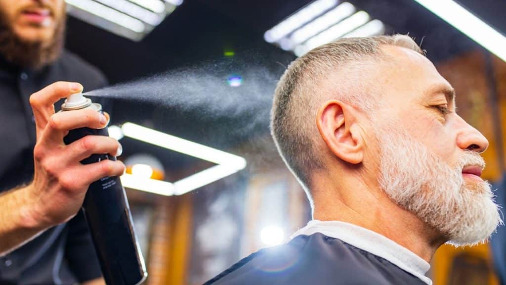A barber spraying hair product on an older man with a white beard in a barbershop.
