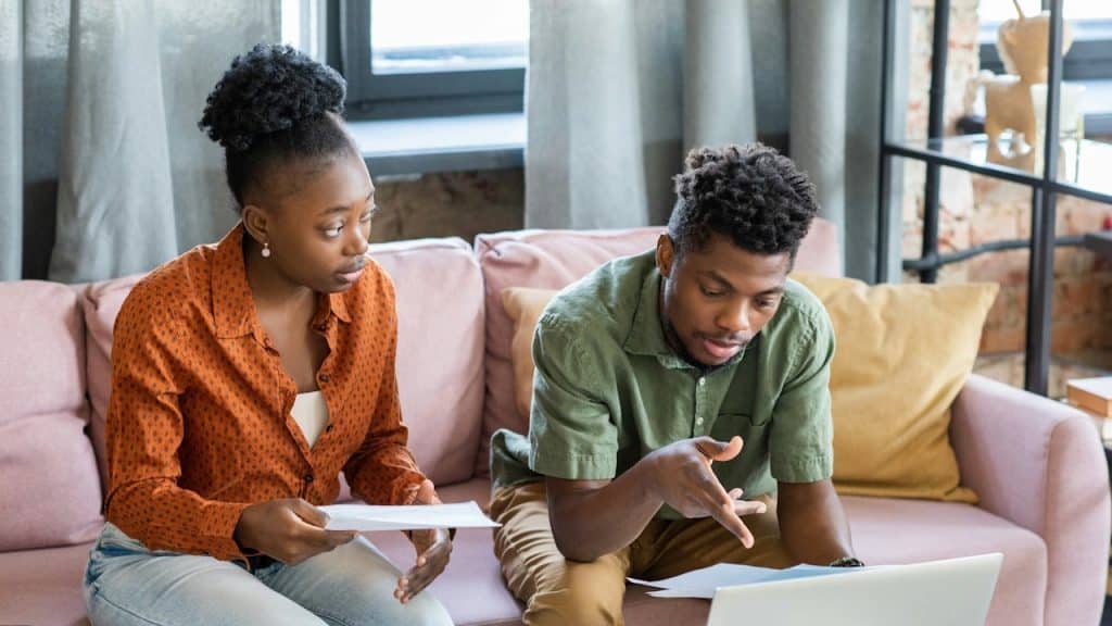Young couple sitting on a pink couch, reviewing documents and looking at a laptop.