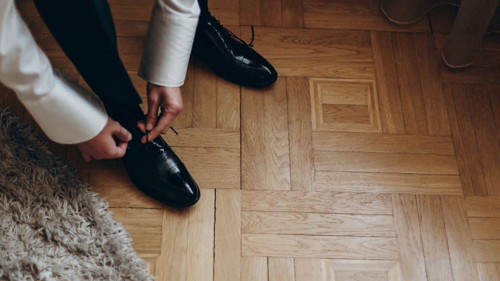 High-angle view of a person wearing a formal white shirt and black trousers, tying the laces of shiny black dress shoes on a parquet floor.