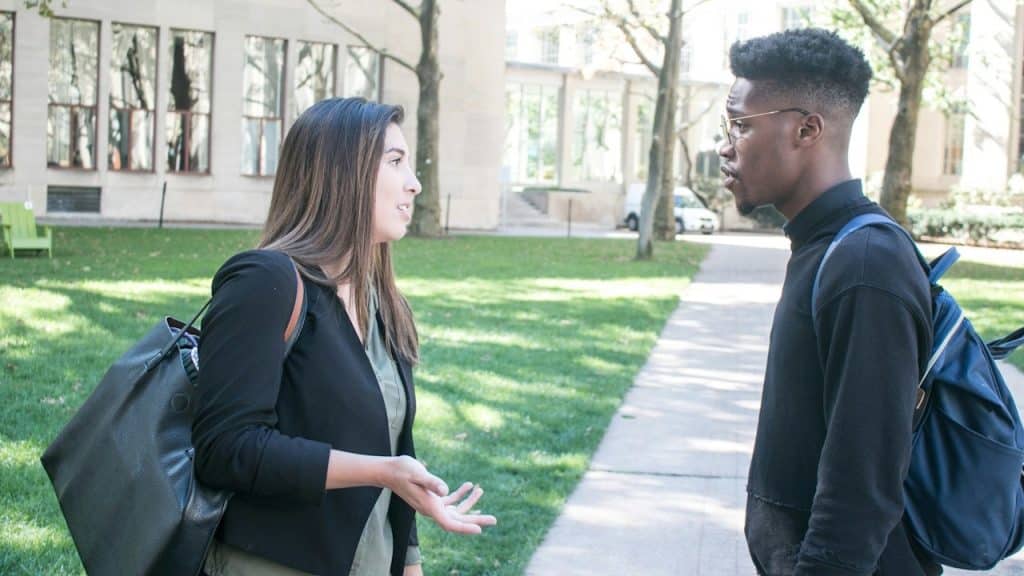 Two college students, a woman and a man with a backpack, having a serious conversation on a university campus lawn.