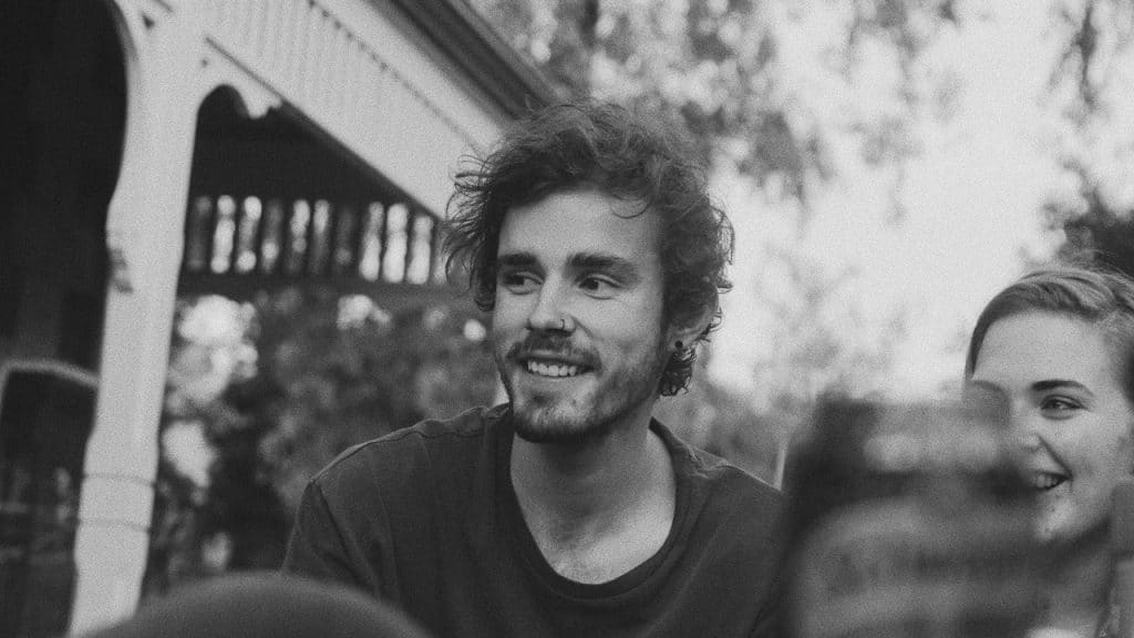 Black and white photo of a smiling young man with curly hair and a mustache, sitting outdoors with a woman beside him.