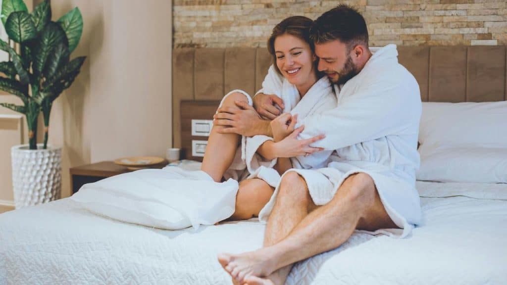 A joyful couple relaxing together in matching bathrobes on a cozy hotel bed.