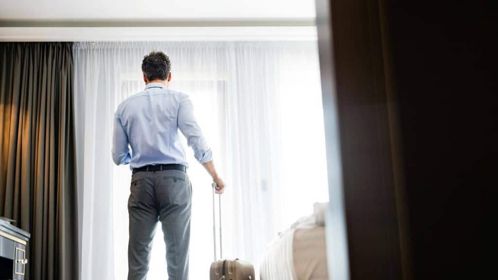 A man gazing out a hotel window, suitcase by his side, deep in thought.