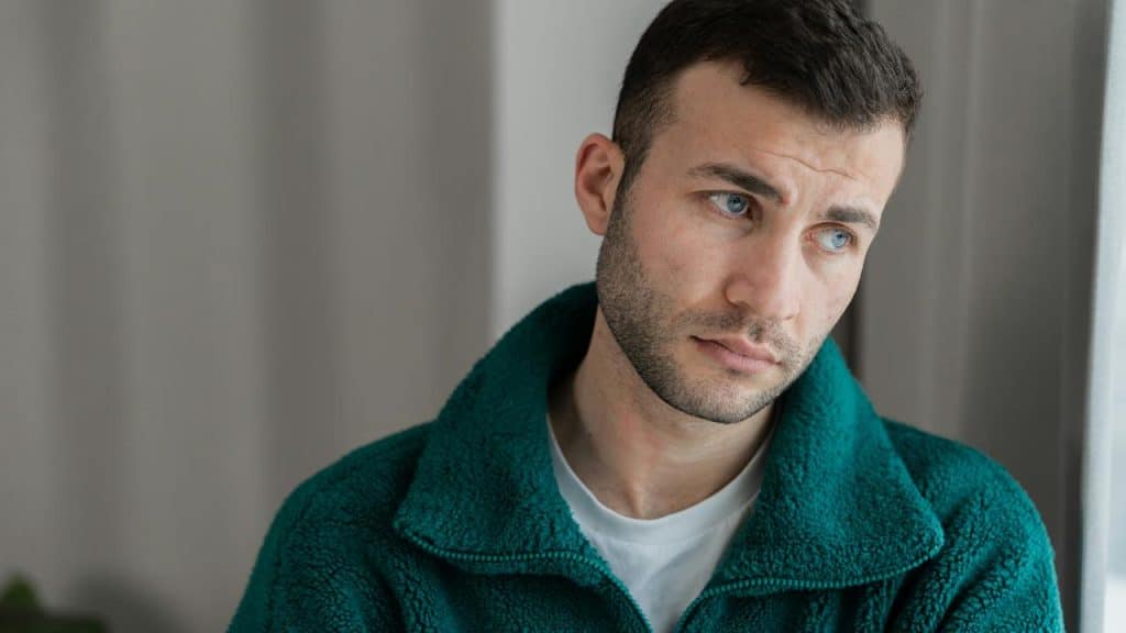 A man in a green jacket staring thoughtfully out a bright window.