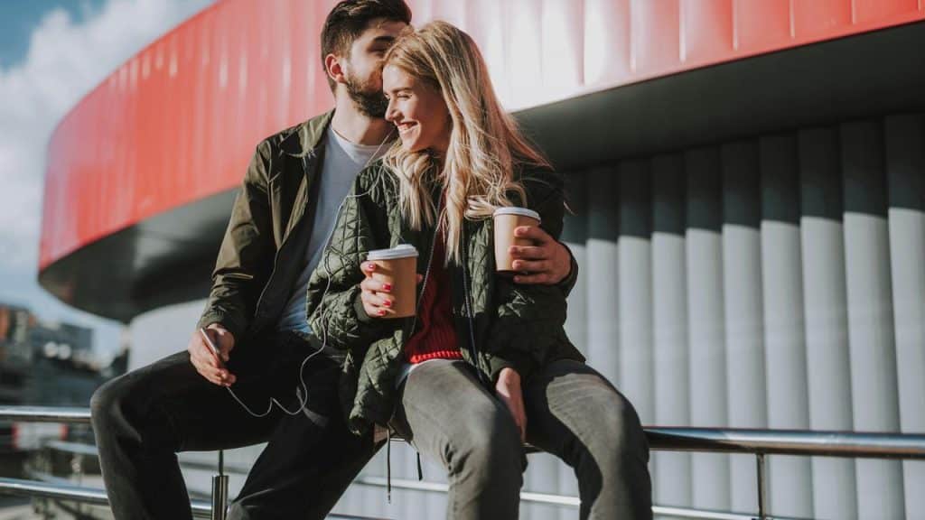 A couple sitting outdoors holding coffee cups and smiling at each other.