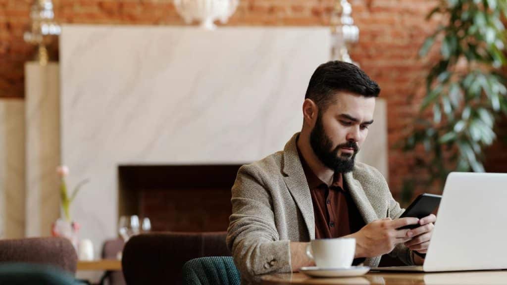 A man sitting in a café, checking his phone beside his laptop and coffee.