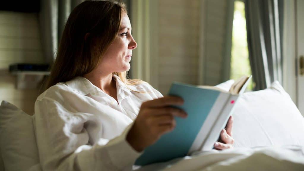 A woman in a white shirt reading a book while sitting in bed.